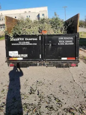 A black dump trailer filled with tree trimmings parked outdoors, displaying contact information for a junk removal service.