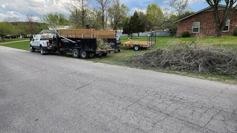 A white truck towing a black dump trailer filled with yard waste, with a smaller flatbed trailer parked on a lawn nearby.
