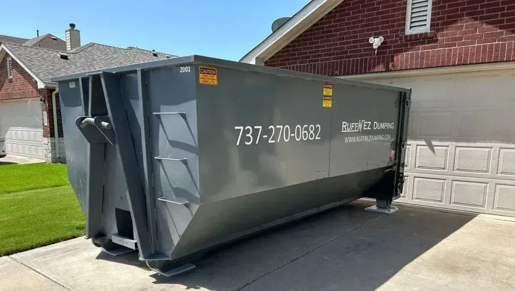 A gray roll-off dumpster parked on a residential concrete driveway in front of a house.