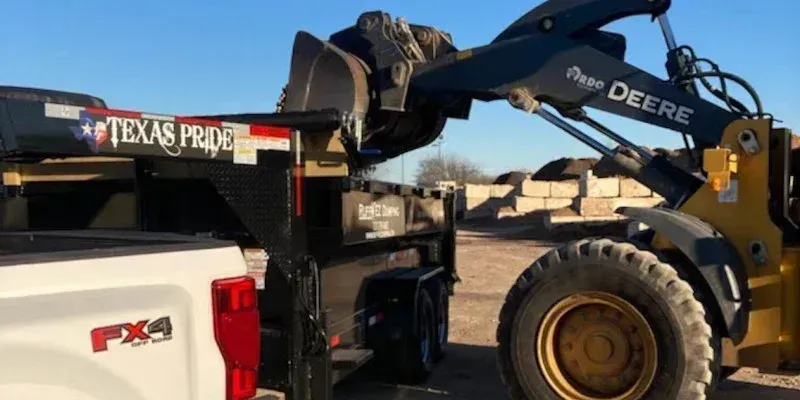 A front loader scoops dirt into a black Texas Pride dump trailer attached to a white pickup truck.
