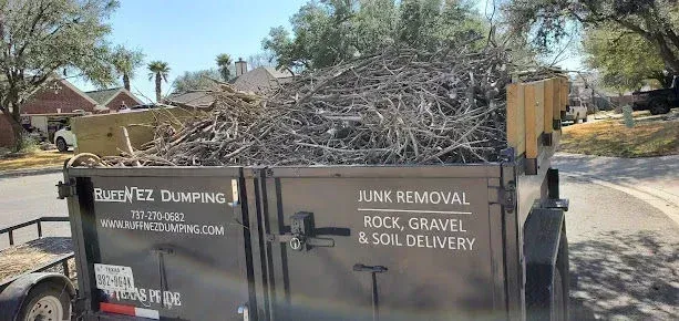 A black dump trailer filled with yard debris parked on a suburban street. 