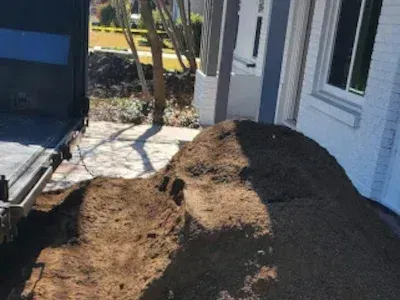 A large pile of dark brown mulch sits on a paved driveway next to a dump truck and a white brick house.