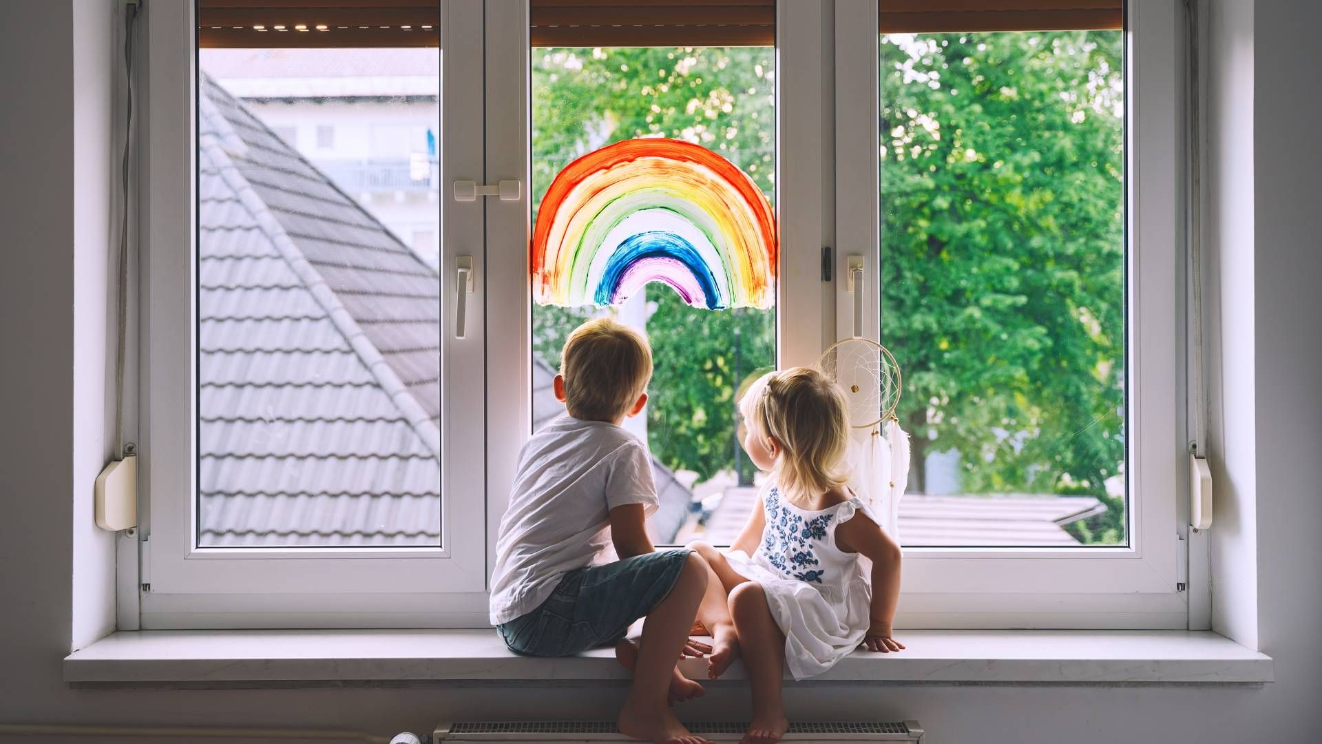 A boy and a girl are sitting on a window sill looking out the window.