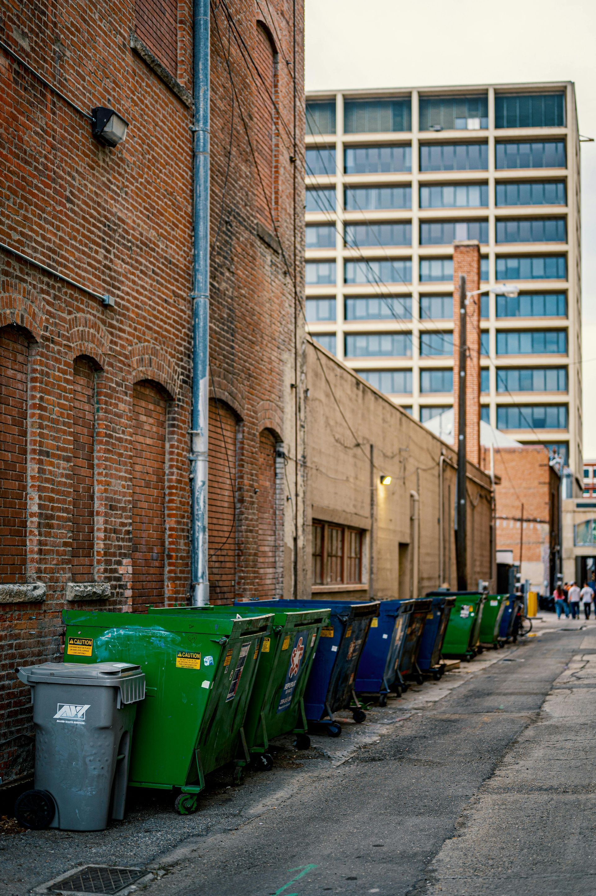 Alleyway with a row of trash bins against brick walls. A modern building is in the background.