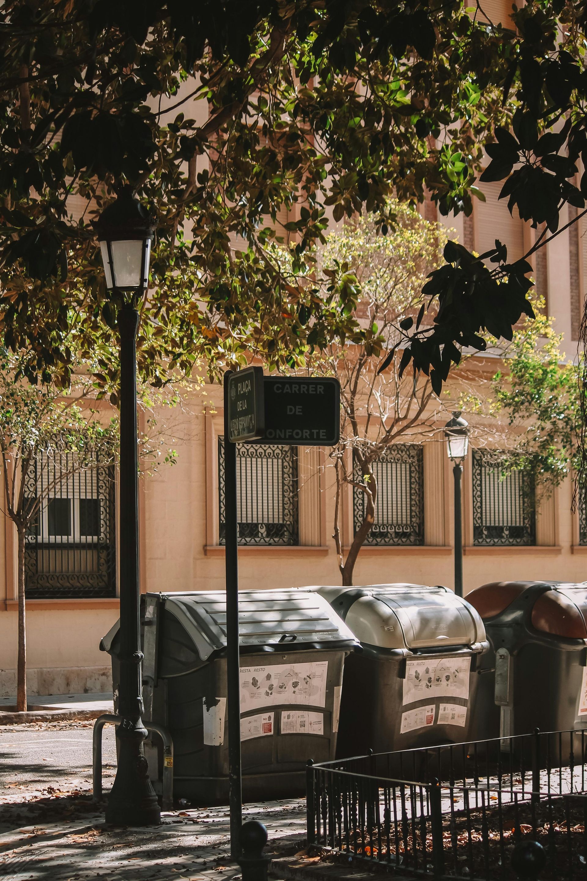 Street scene with trash bins, streetlights, and building. Sunlight filters through trees.