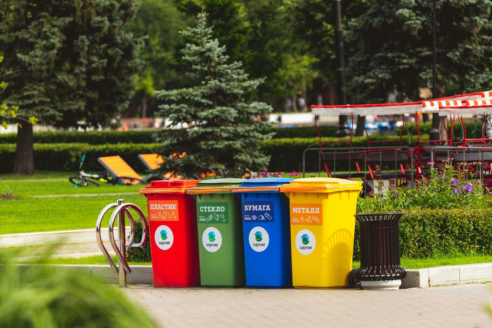 Four colorful recycling bins in a park: red, green, blue, yellow; trees and greenery in the background.