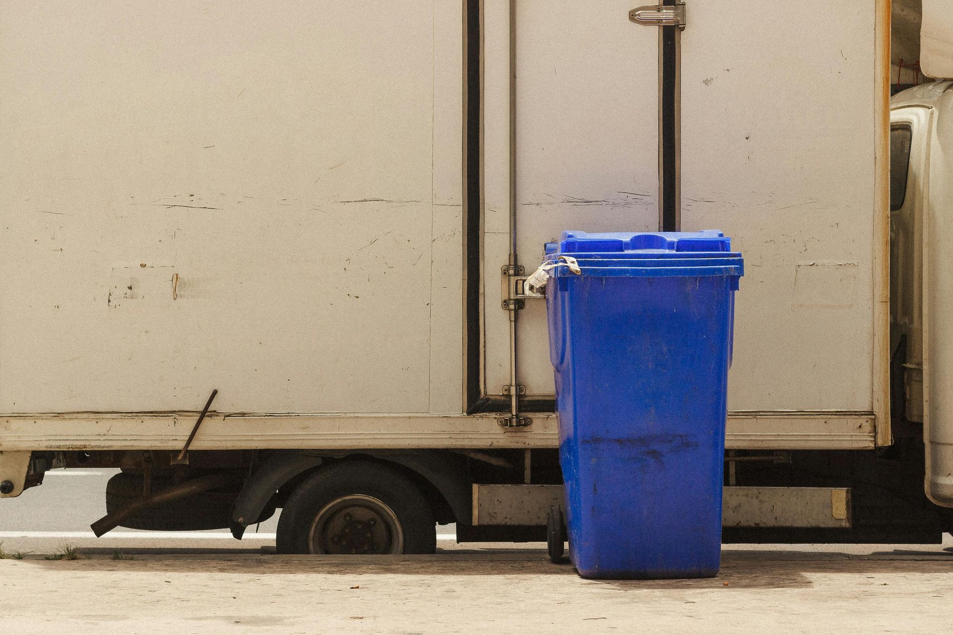 Blue trash bin next to a white truck, parked on a concrete surface.