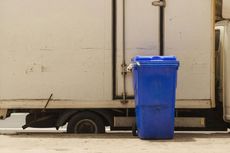 Blue trash bin next to a white truck, parked on a concrete surface.