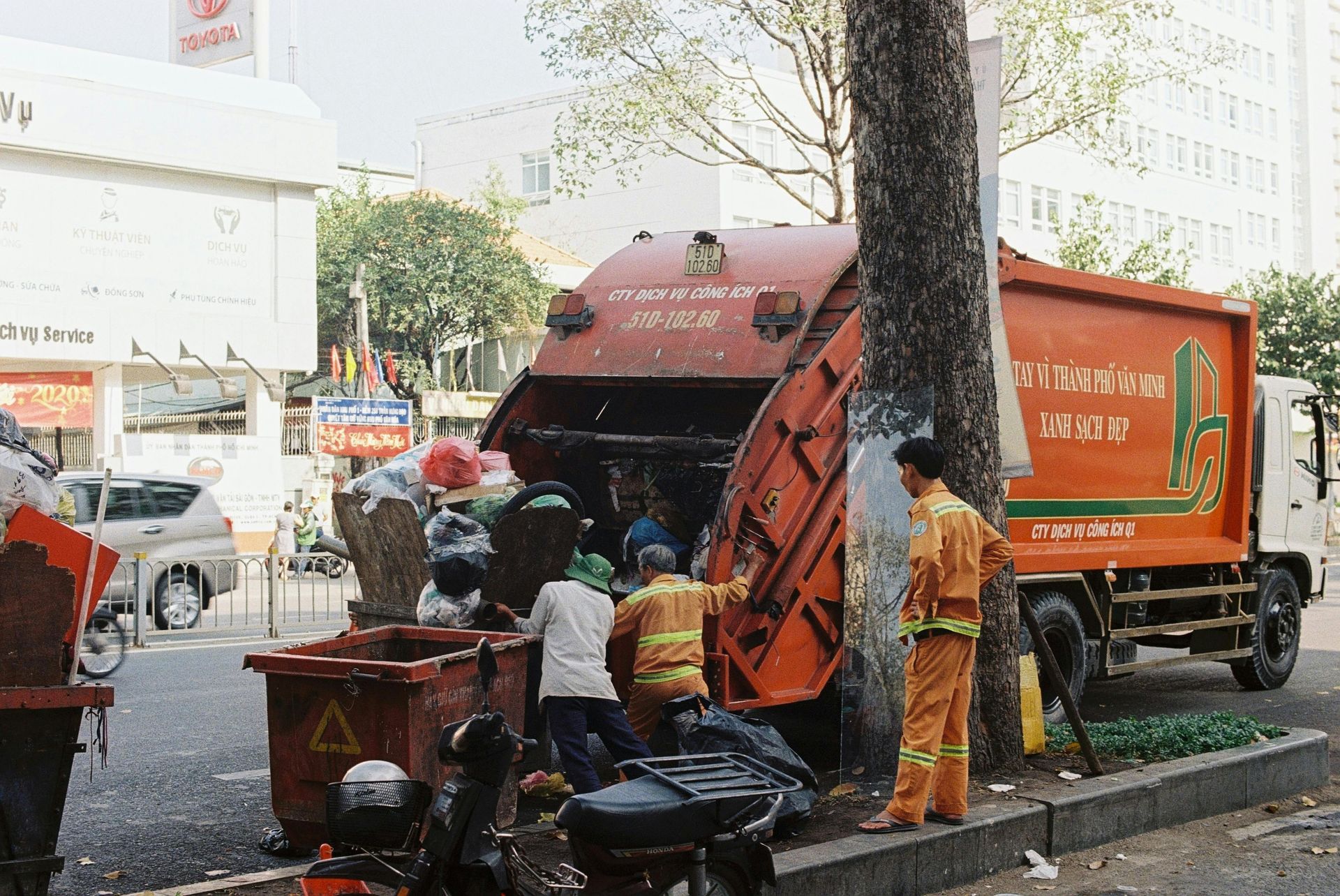 Garbage truck loading trash from a bin on a city street, workers in orange uniforms, tree, buildings.