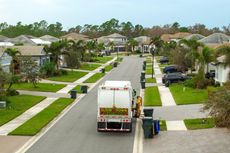 A garbage truck collecting trash bins on a residential street. Homes with green lawns line the road.