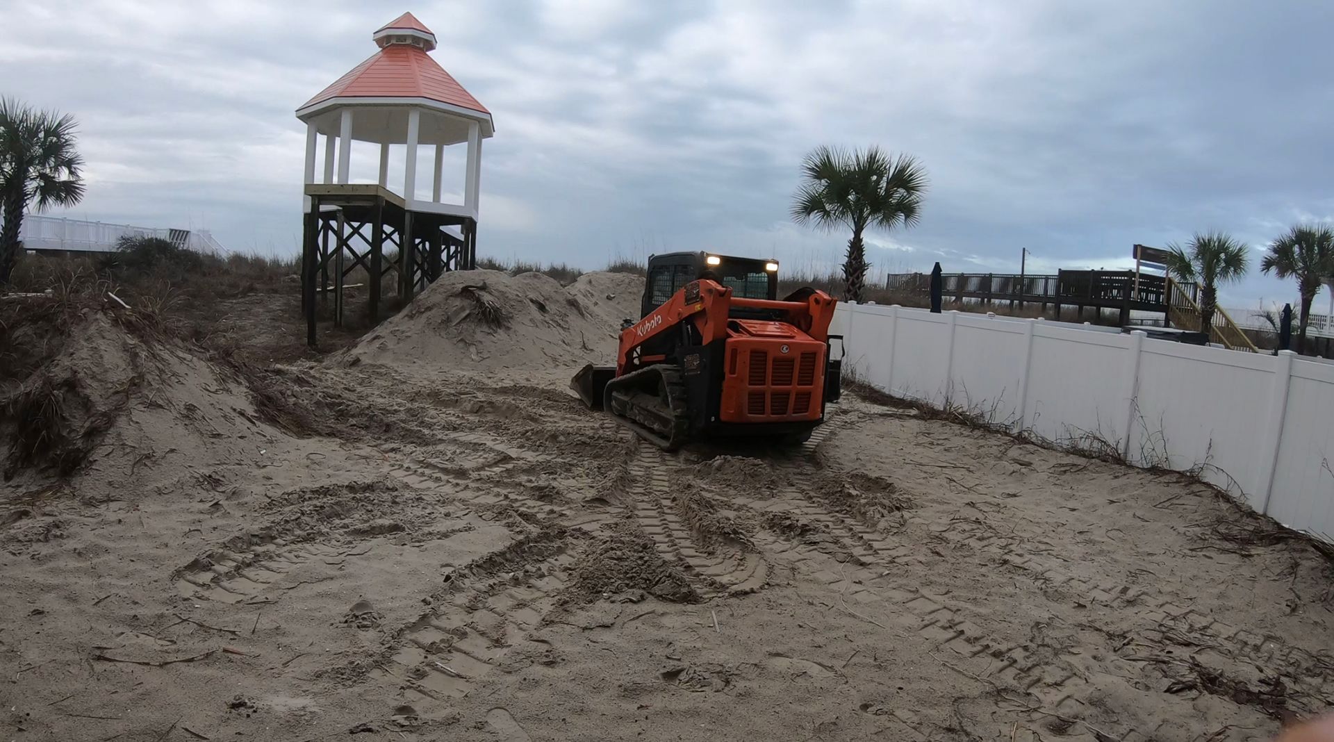 A bulldozer is digging a hole in the sand near a gazebo.
