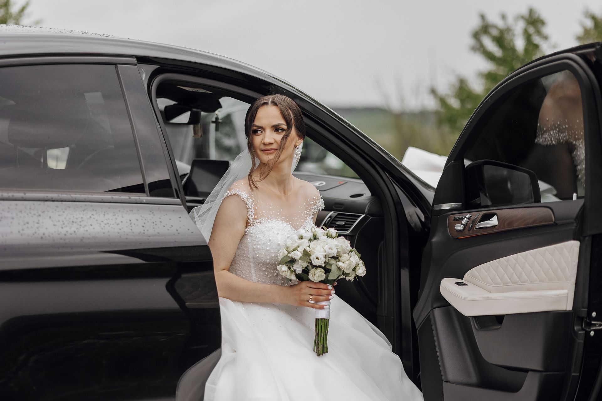 Bride in a white wedding dress holding a bouquet beside an open car door, looking at the camera.