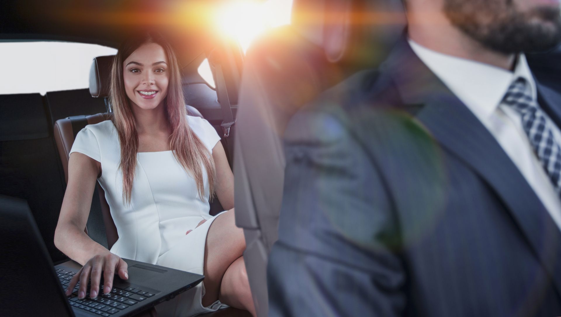 Smiling woman in a white dress sitting in a car, using a laptop in bright sunlight