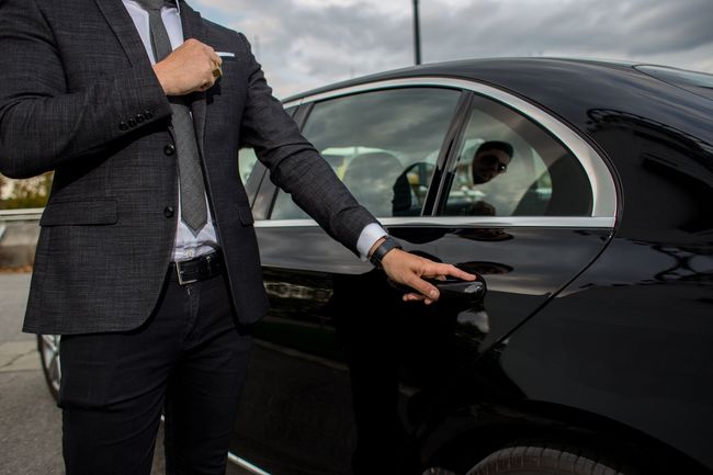 Man in a gray suit opening a black car door outdoors under a cloudy sky