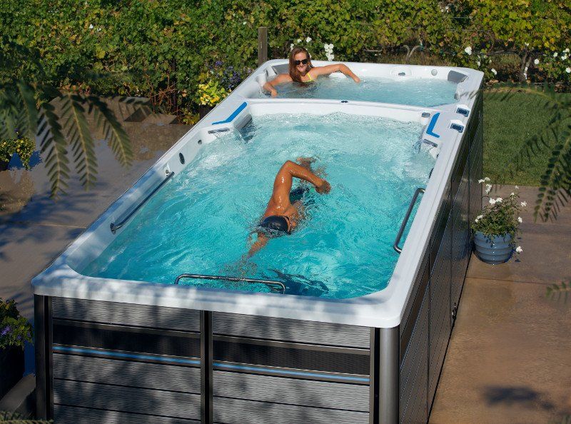 Two people in a swim spa. One swims freestyle, other relaxes. Outdoor setting.