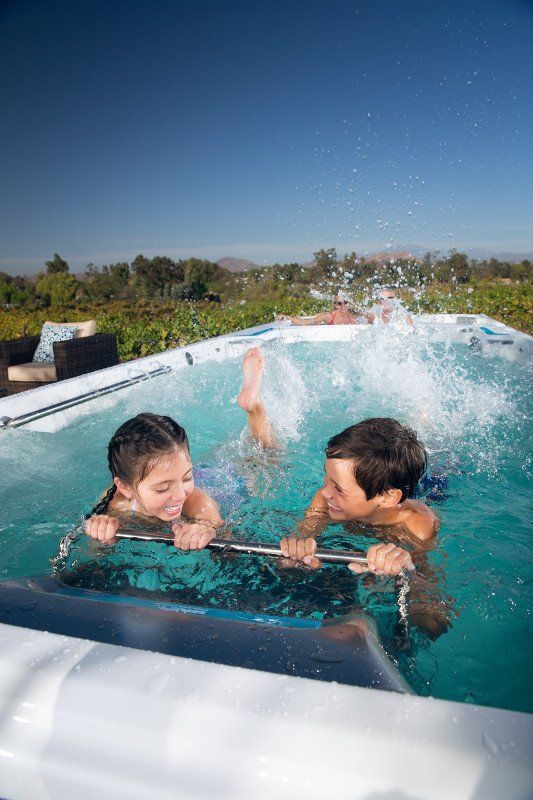 Two kids smile, playing in a blue, rectangular pool; water splashes up as one kicks.