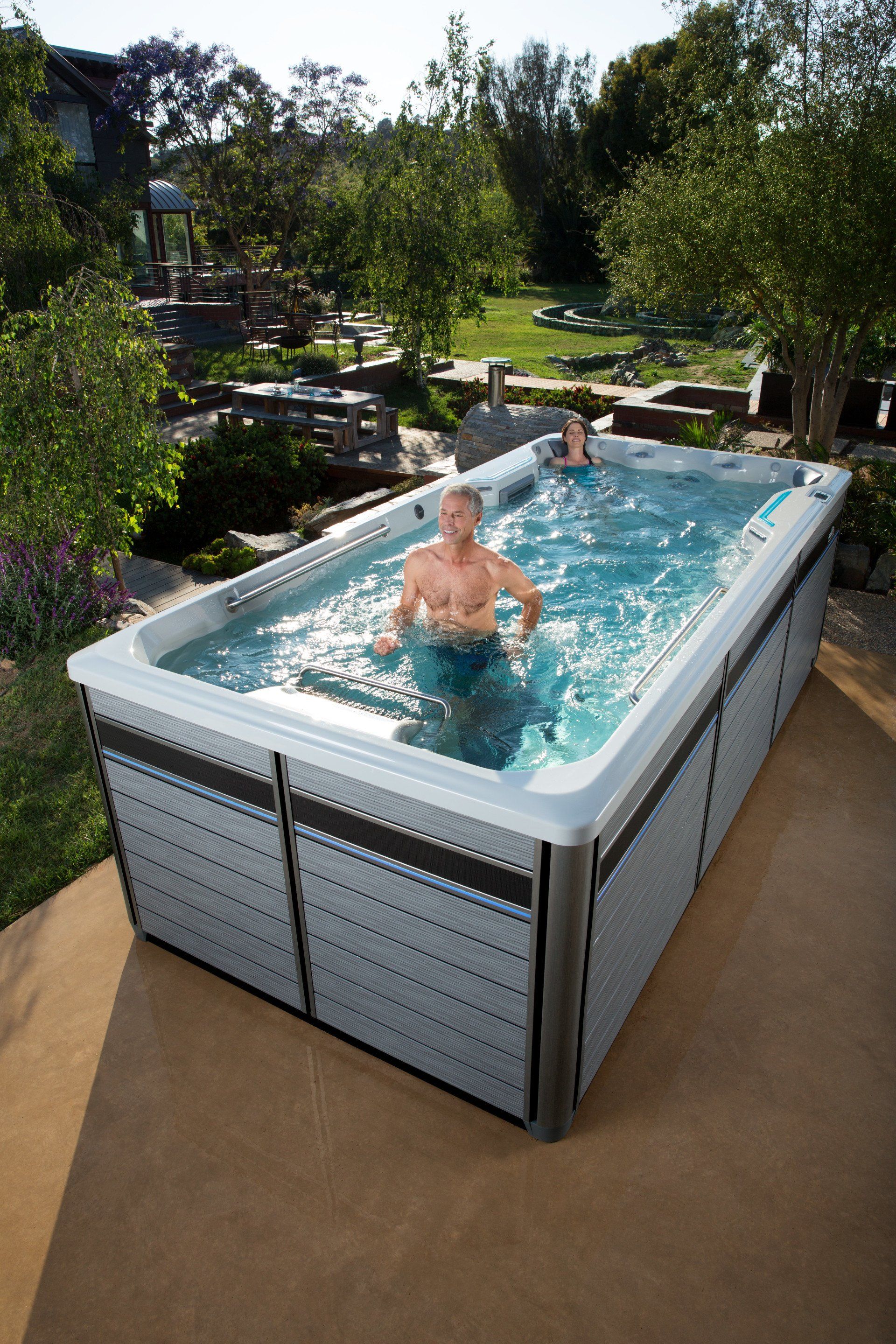 Man and woman in a rectangular swim spa outdoors. Gray panels surround the spa. Lush greenery in the background.