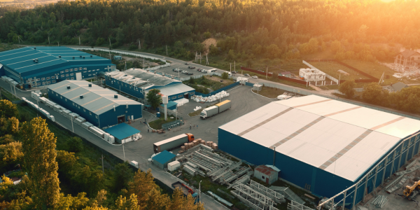 Aerial view of an industrial complex with blue-roofed buildings, a white-roofed warehouse, and trucks in a forested area.