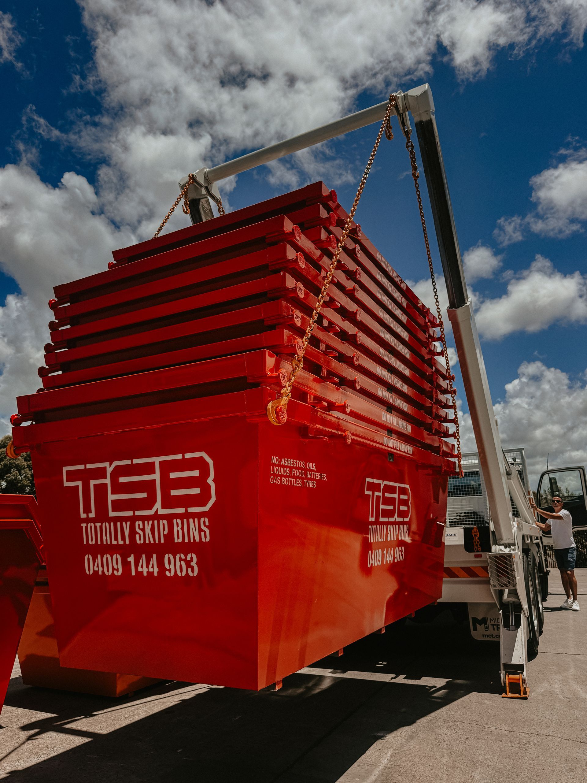 Red Dumpster Being Lifted By A Crane On A Trailer, Under A Blue Sky. Text On Dumpster Says 