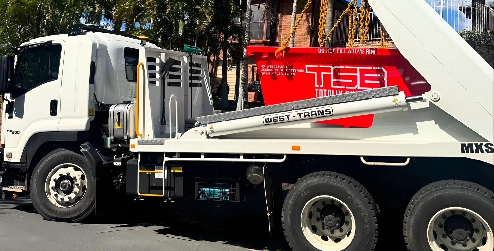 White skip bin truck lifting a bright red waste container labeled  on a residential street