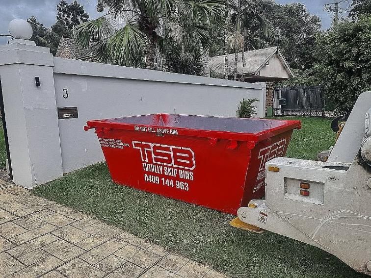 A Red Dumpster is Sitting in the Grass Next to a White Wall — Totally Skip Bins in Gold Coast, QLD