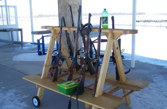 Rifles on a wooden rack at an outdoor shooting range. A green bottle and a small green box are also present.