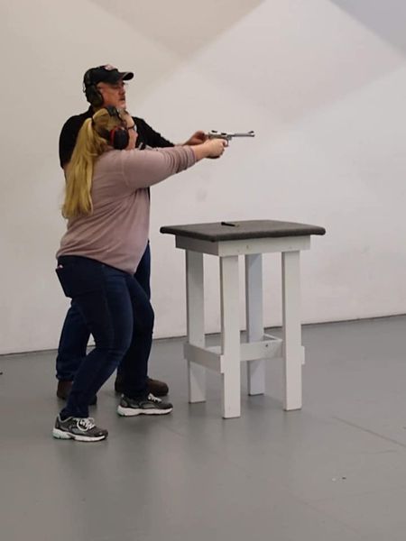 Instructor guides a woman aiming a gun at a shooting range. Both wear ear protection.