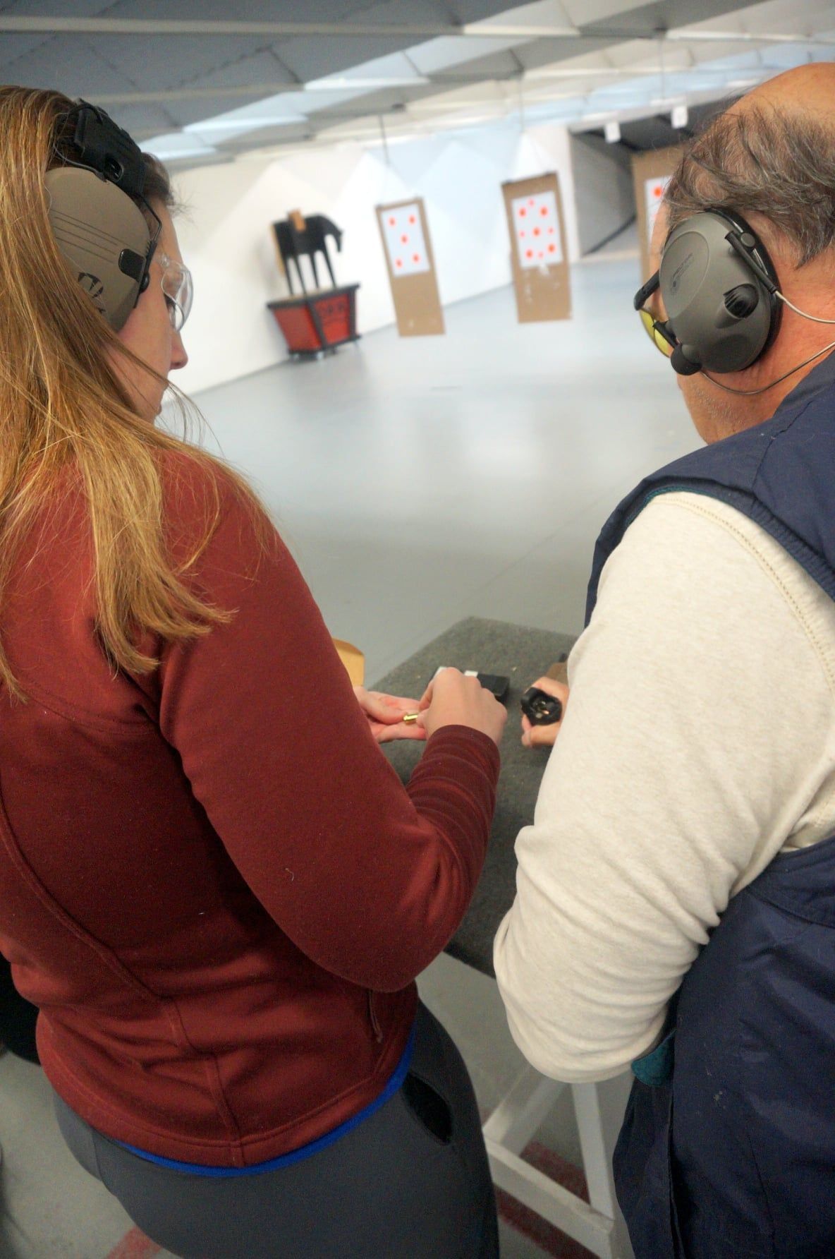 Woman and man at a shooting range. She's holding a gun, both wearing ear protection. Targets visible.
