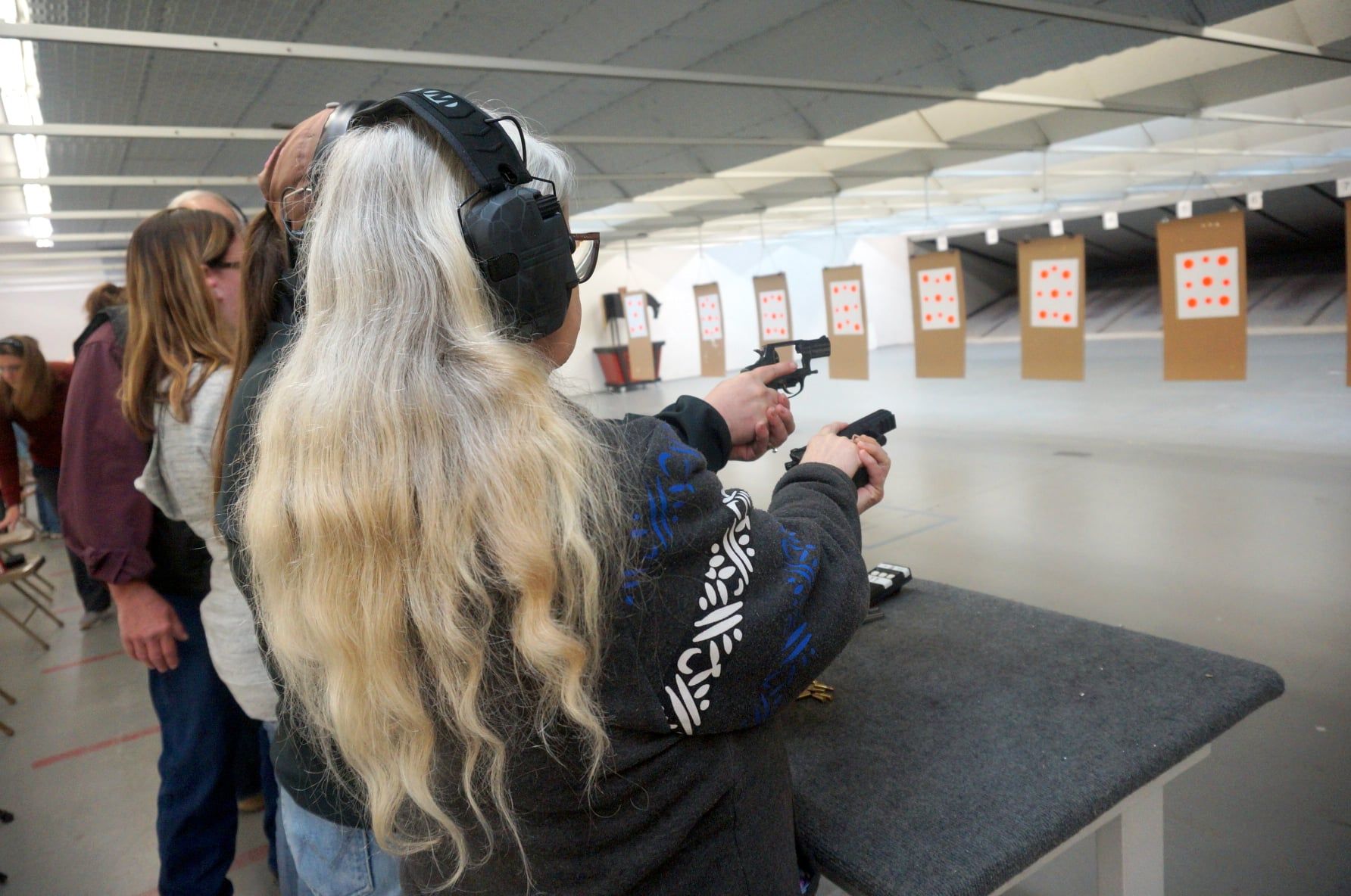 Woman with long, blonde hair shoots a handgun at a target in a shooting range.