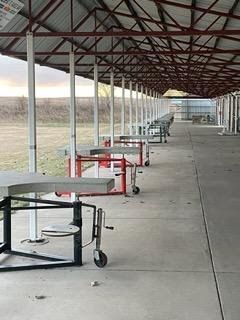 Shooting range with covered stalls, concrete floor, and tables. Metal roof, white support poles, and a field in the background.
