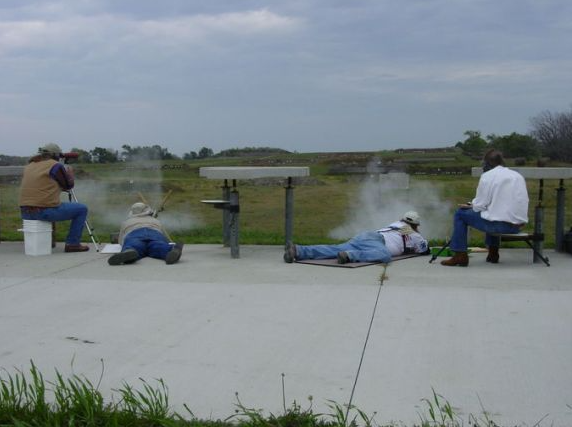 Four people firing rifles at a shooting range; smoke rising from the guns, concrete pad, rural setting.