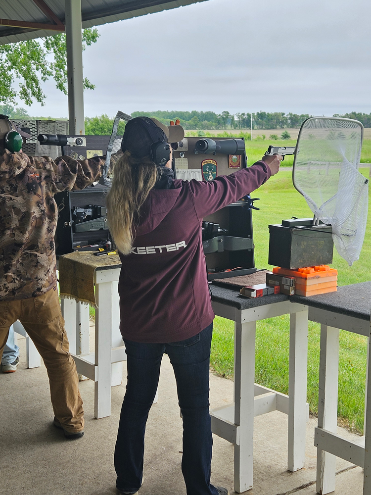 Woman shoots a handgun at an outdoor shooting range, wearing a maroon jacket and ear protection.