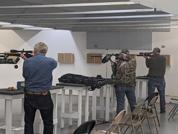Three people aiming rifles at a shooting range, indoors. Beige walls, white tables, and chairs are visible.