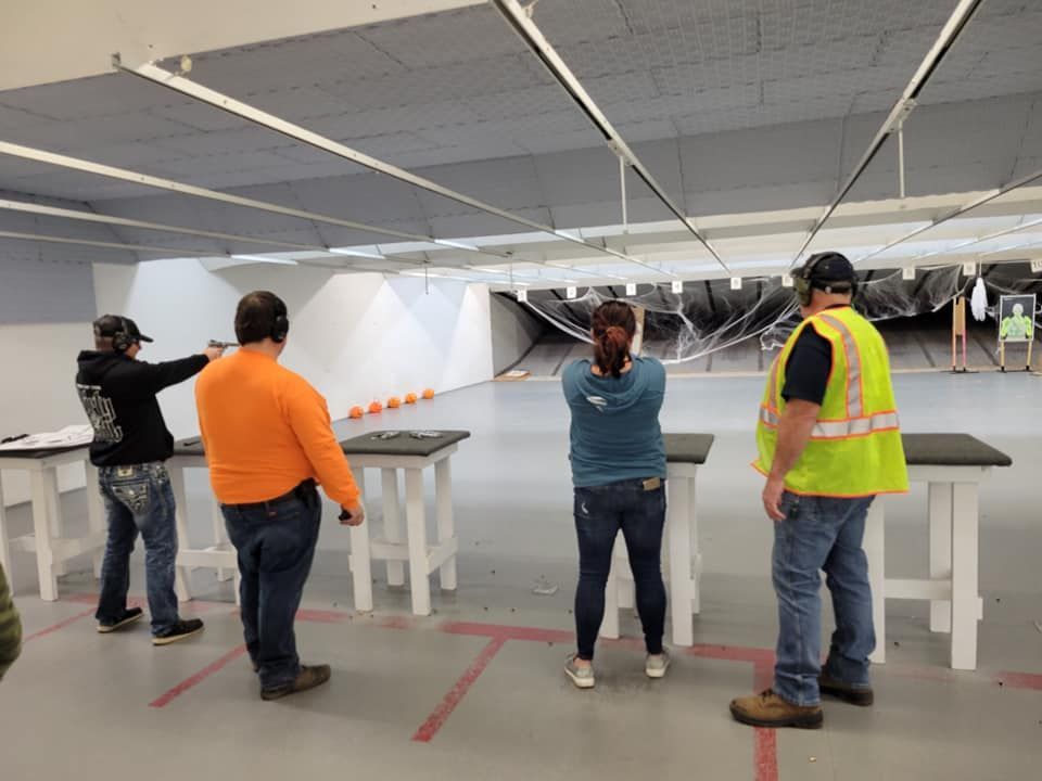 People shooting guns at a range; one in orange shirt, one in green safety vest.