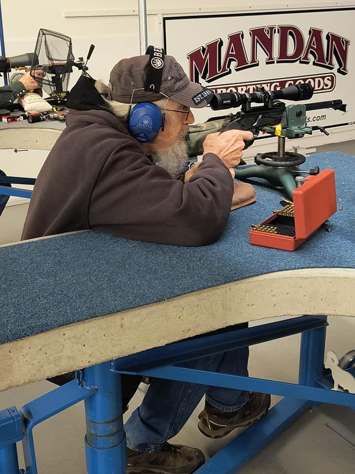Man in earmuffs and hat, aiming a rifle at a shooting range, with a box of bullets nearby.