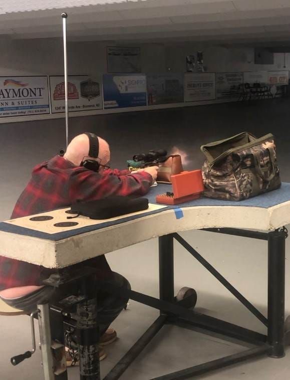 Man in plaid shirt shooting a rifle at a range, wearing ear protection.