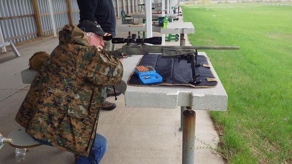 Person in camouflage jacket aiming rifle at a shooting range.