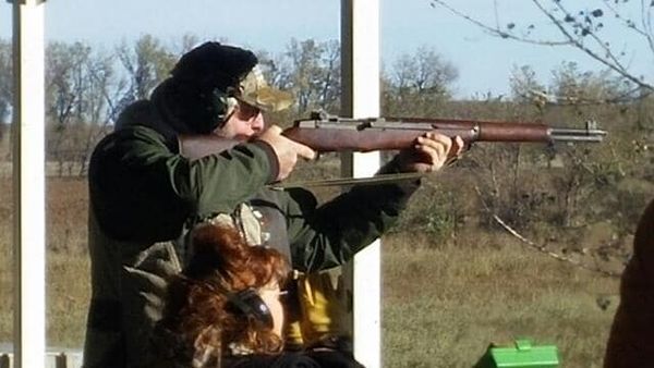 Man wearing ear protection aims a rifle at an outdoor shooting range.