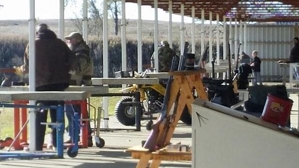 People at an outdoor shooting range with rifles, a yellow motorcycle, and a wooden structure.