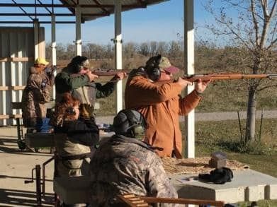 People aiming rifles at an outdoor shooting range, wearing ear and eye protection.