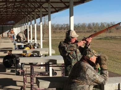 Men in camouflage shoot rifles at an outdoor shooting range under a covered shelter.