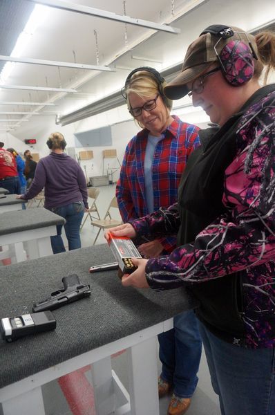 Two women at a shooting range, one loading bullets, wearing ear and eye protection.