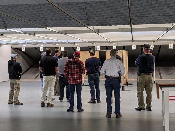 Group of people at an indoor shooting range observing targets; some are wearing tactical gear.