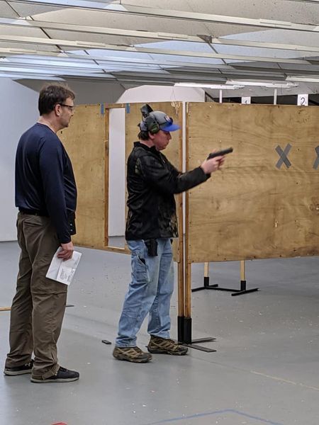 Man aims a handgun at a target in a shooting range, observed by another man.