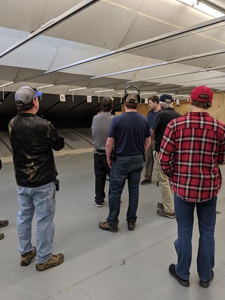 Group of people at a shooting range, wearing safety glasses, focused on targets in the distance.