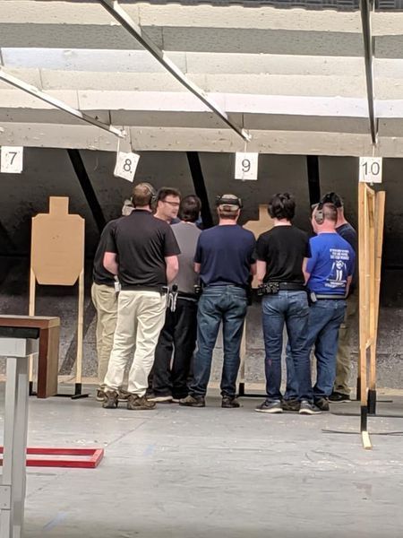 People standing at a shooting range, looking at targets. Beige, gray, and blue attire. Numbers 7, 8, 9, and 10 above the targets.