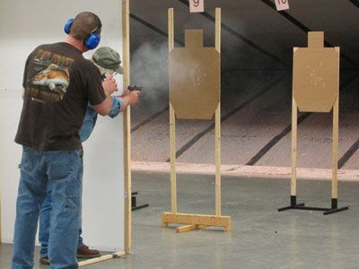 Man firing handgun at target in a shooting range; another man observing. Smoke visible.