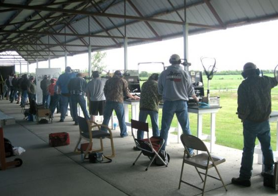 People shooting at a gun range outdoors. Several people wear ear protection and face targets.