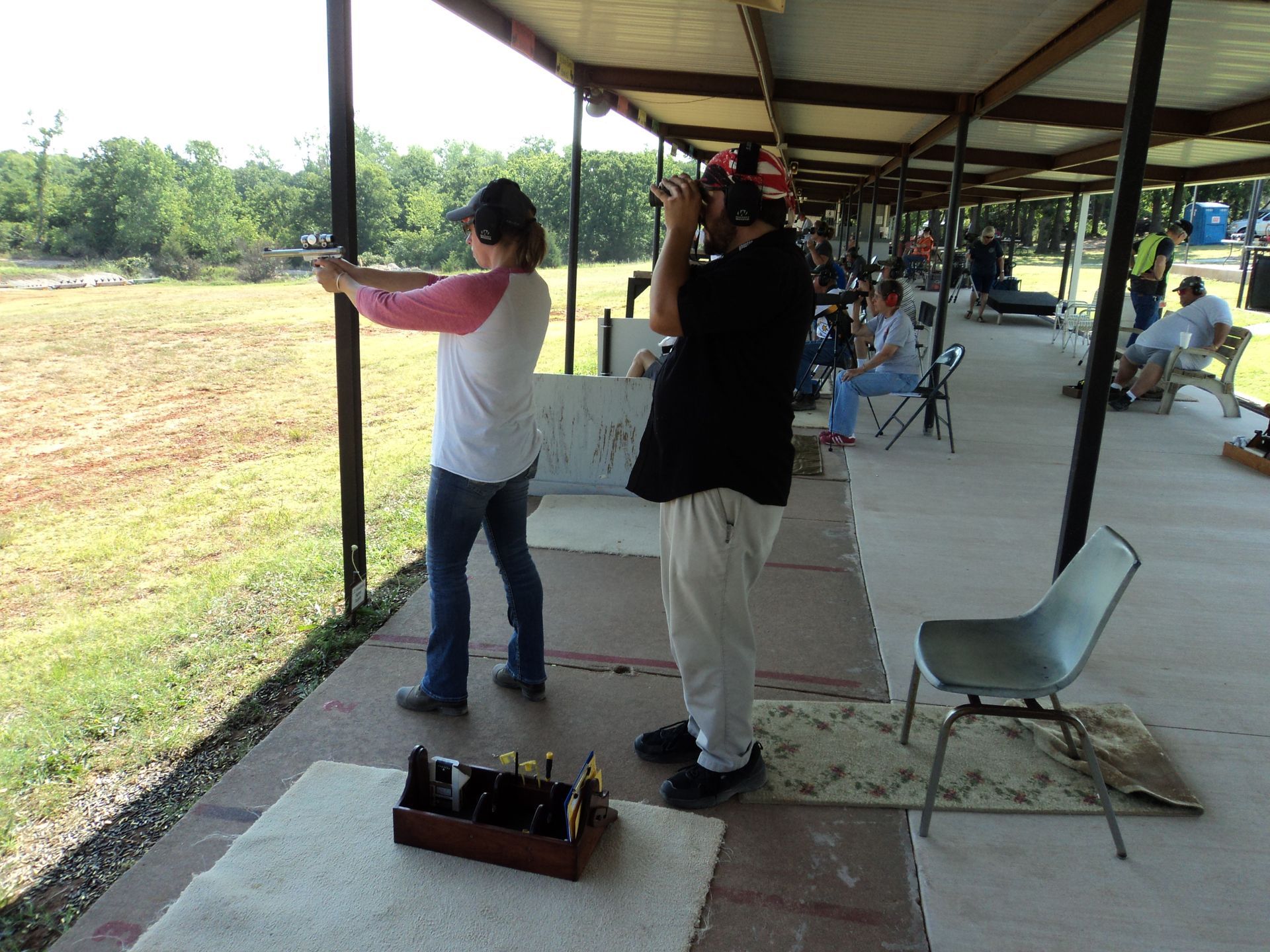 Woman aiming a gun at a shooting range, man observing. Outdoors, sunny.