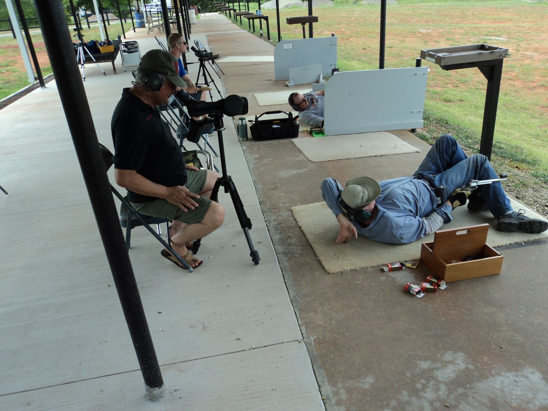 People at an outdoor shooting range. One sits with a scope, another lies down. Concrete pad, green field.